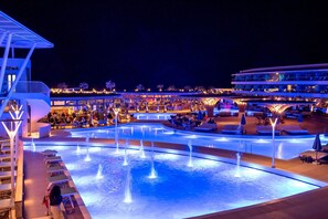 Un espace piscine d’un complexe hôtelier, aménagé pour la nuit, avec plusieurs niveaux de terrasses et de coins repas, le tout entouré de palmiers.