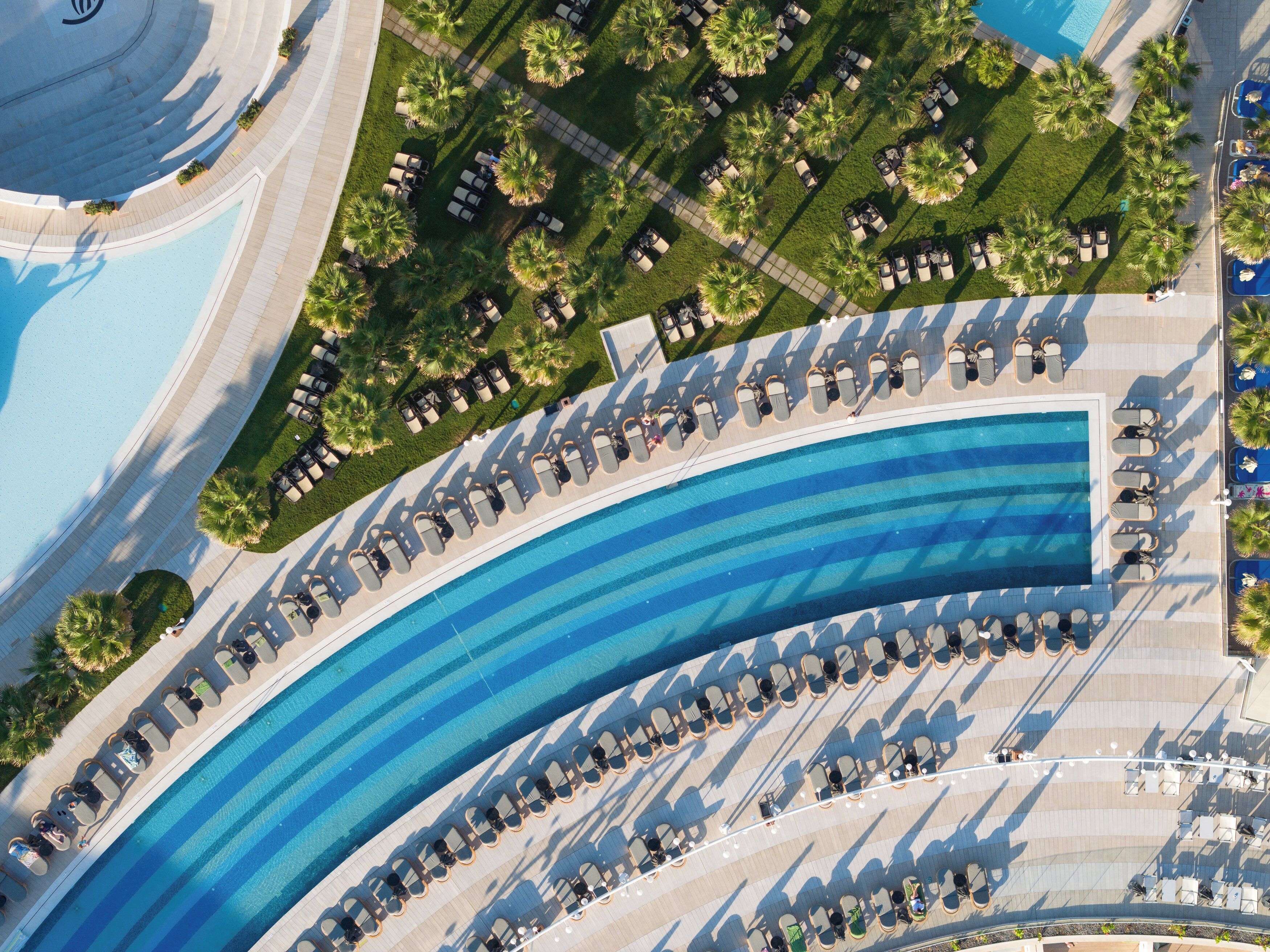 An aerial view of a hotel with a large curved pool, sun loungers, and palm trees.