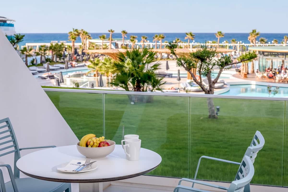 A balcony with a table set for breakfast, overlooking a pool area with palm trees and a view of the ocean.