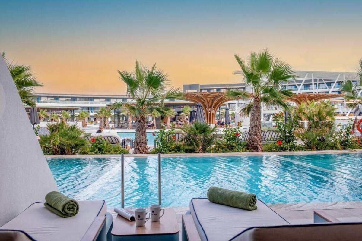 A poolside area with lounge chairs, a table with a towel and a cup, and a view of a modern building and palm trees.