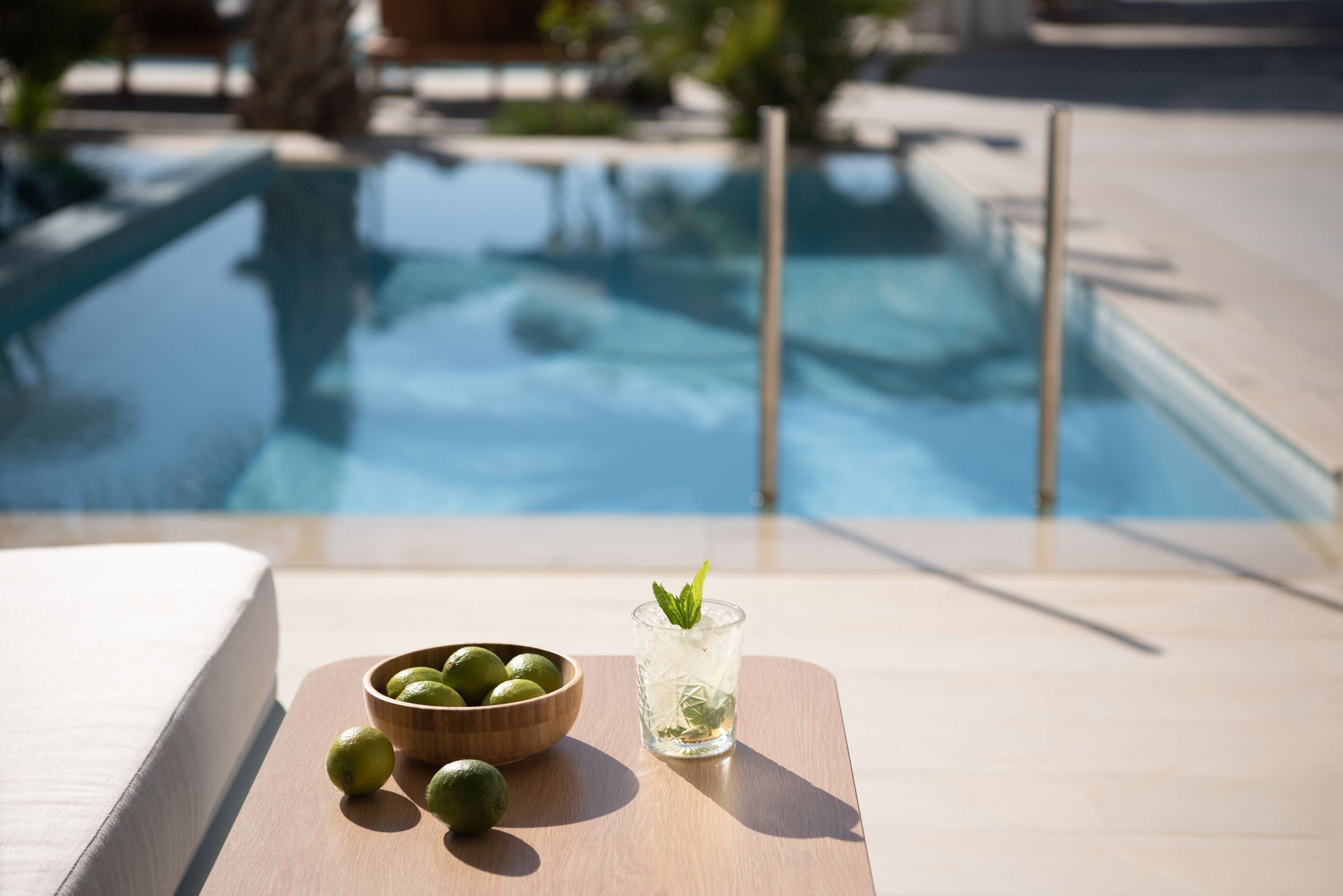 A poolside setting with a wooden table holding a glass of drink and a bowl of limes.