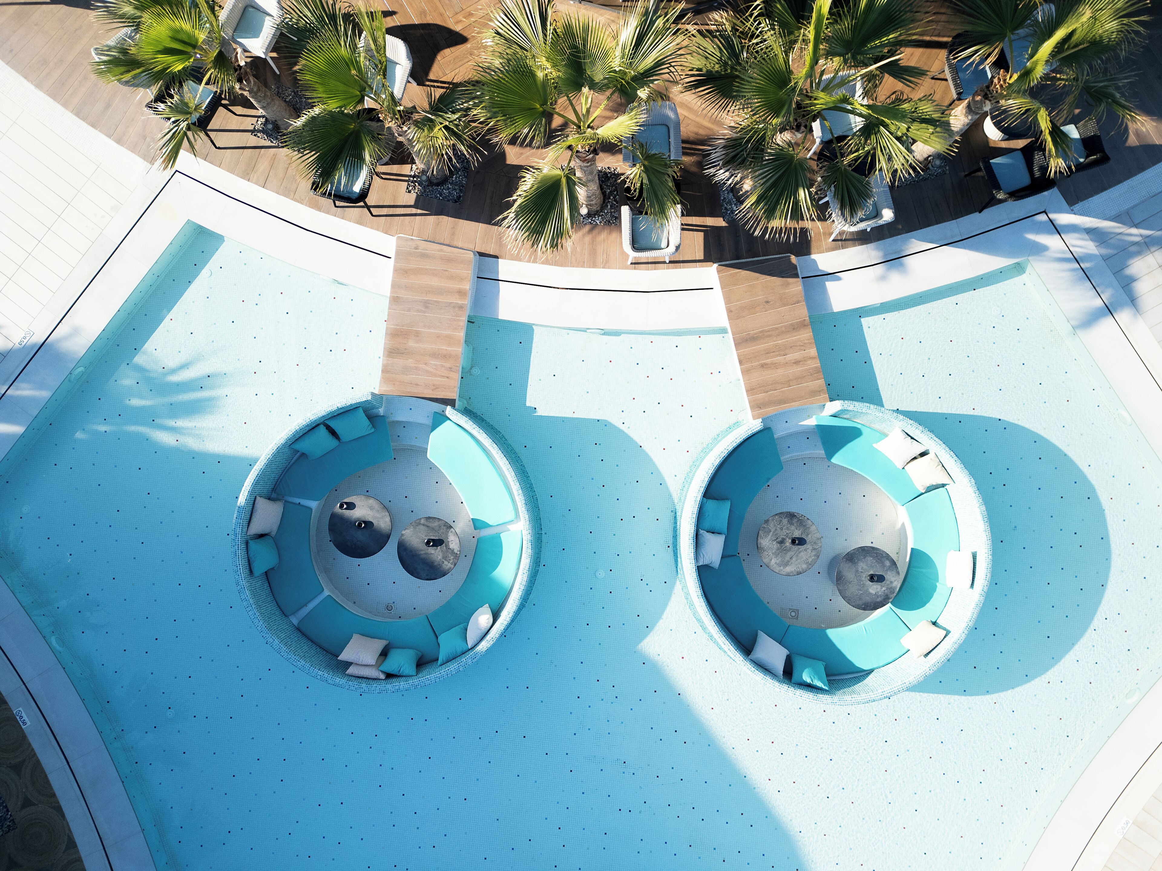 Vista aérea de un área de piscina con dos fuentes circulares, sillas de descanso y palmeras.