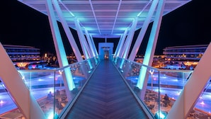 Un puente moderno con barandas de vidrio e iluminación azul, que conduce a una zona de piscina con mobiliario exterior.