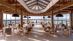 A spacious dining area with multiple tables and chairs, large windows offering a view of the ocean, and a wooden ceiling structure. - Lyttos Mare Adults Friendly 12+ (Hersonissos)