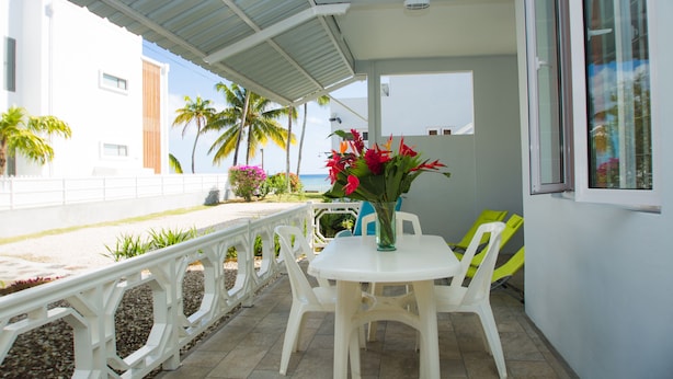 Un portico coperto con tavolo e sedie bianche, un vaso di fiori e vista sulla spiaggia e palme.
