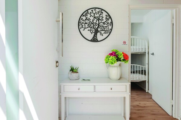 A white room with a desk, a wall-mounted tree decoration, a small potted plant, and a vase with flowers.