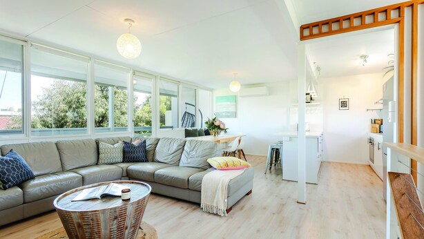 A modern living room with a grey sectional sofa, a round wicker coffee table, and a kitchen area with white cabinets and a wooden countertop. - Aldinga Beach Breakaway - Coastal Beauty Perfect For a Fam Vacay ~ C21 (Aldinga Beach)
