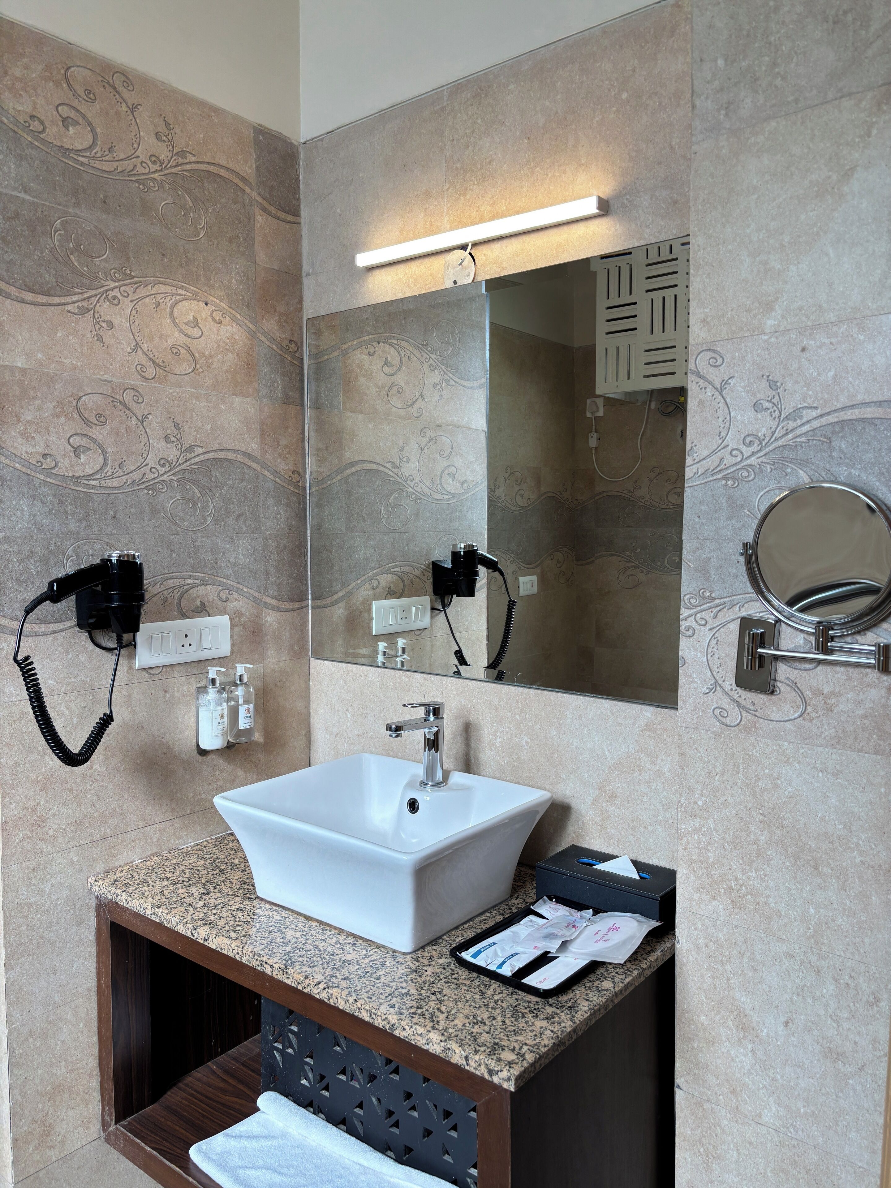 Un baño moderno con un lavamanos blanco, encimera de granito y un espejo grande. Hay dos botellas de champú y acondicionador en la pared, y un perchero con una toalla.