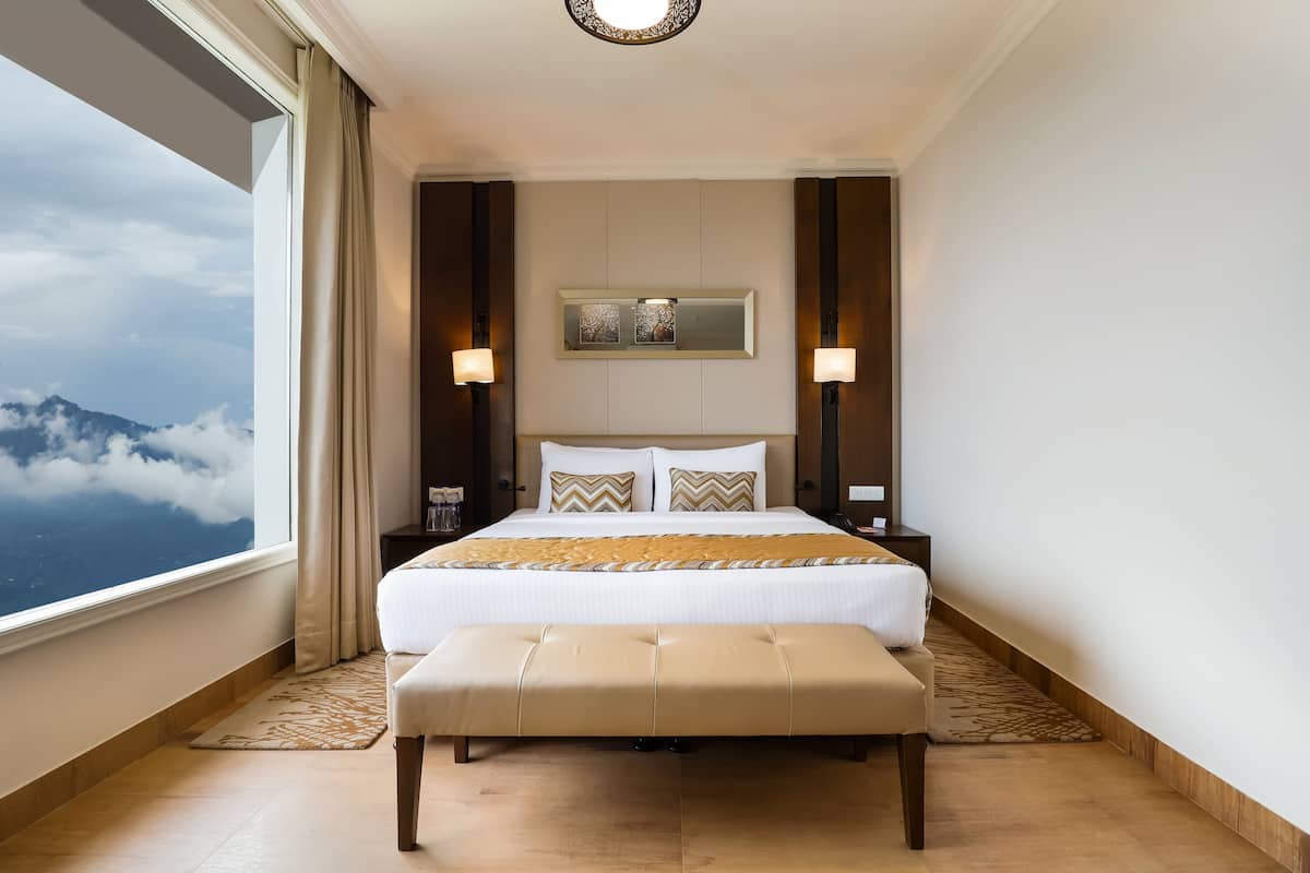 A hotel room with a large bed, two bedside tables, a bench, a large window with a view of clouds and mountains, and a ceiling light fixture.