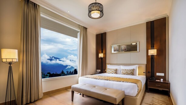 A hotel room with a large bed, a bench, a floor lamp, a nightstand, and a view of the mountains. - Regenta Resort & Spa Mashobra (Shimla)