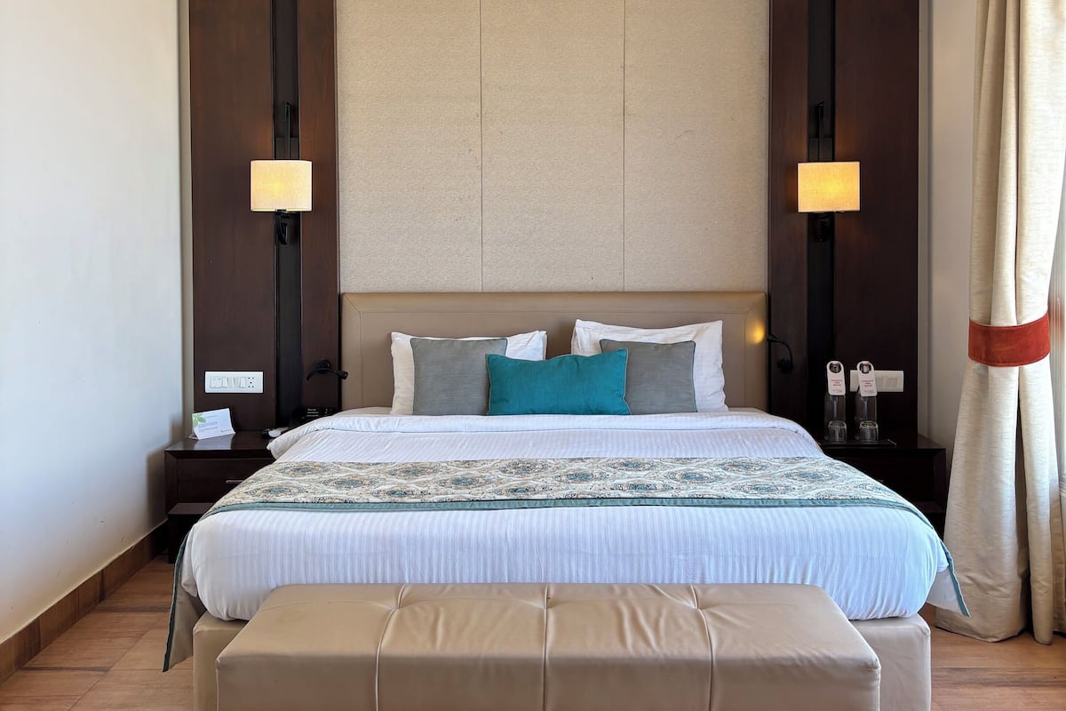 A hotel room with a large bed, a bench, two bedside tables with lamps, a decorative ceiling light, and a large window with curtains.