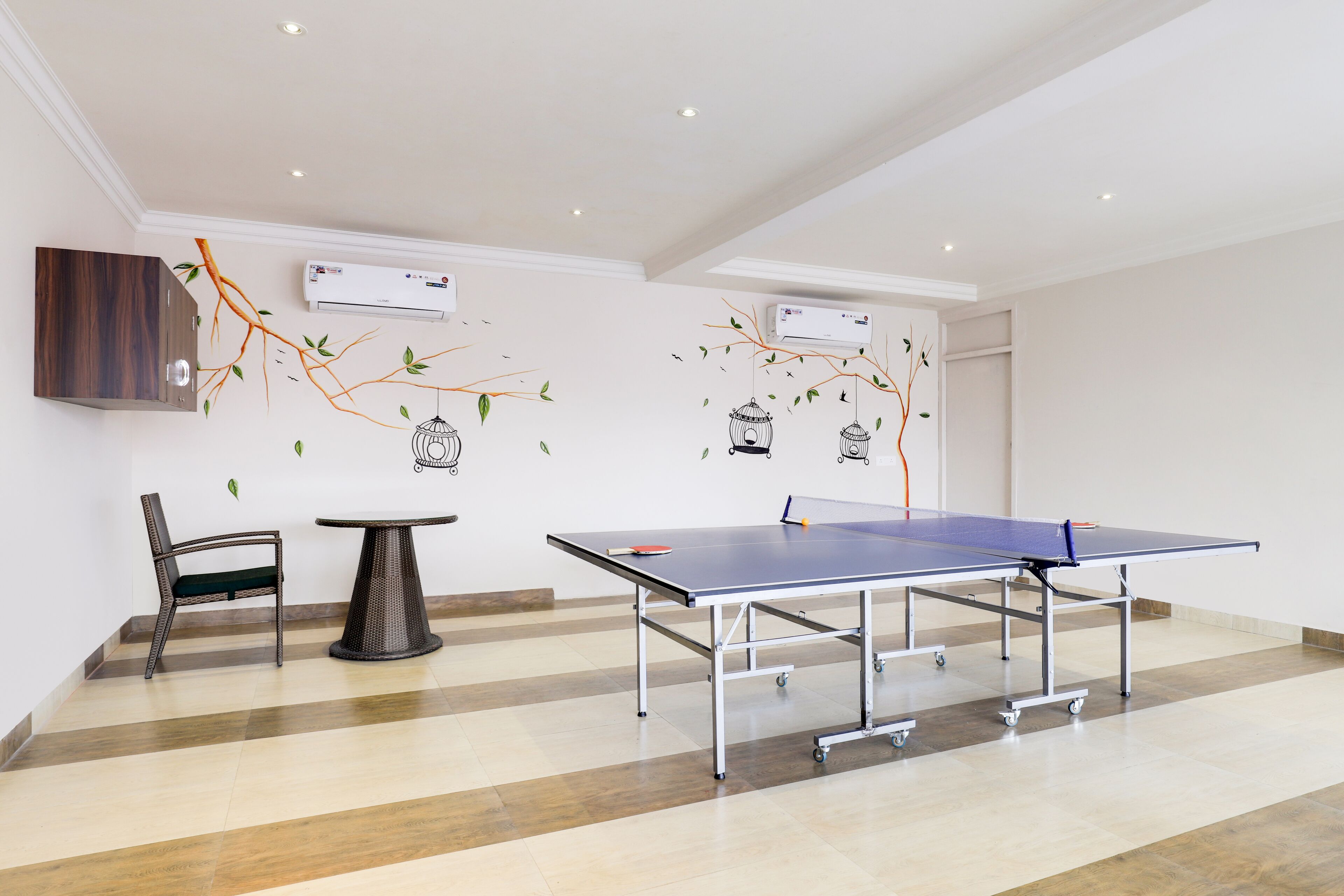 Una mesa de ping pong en una habitación con piso de madera, una silla y adornos en la pared que incluyen ramas y pájaros.