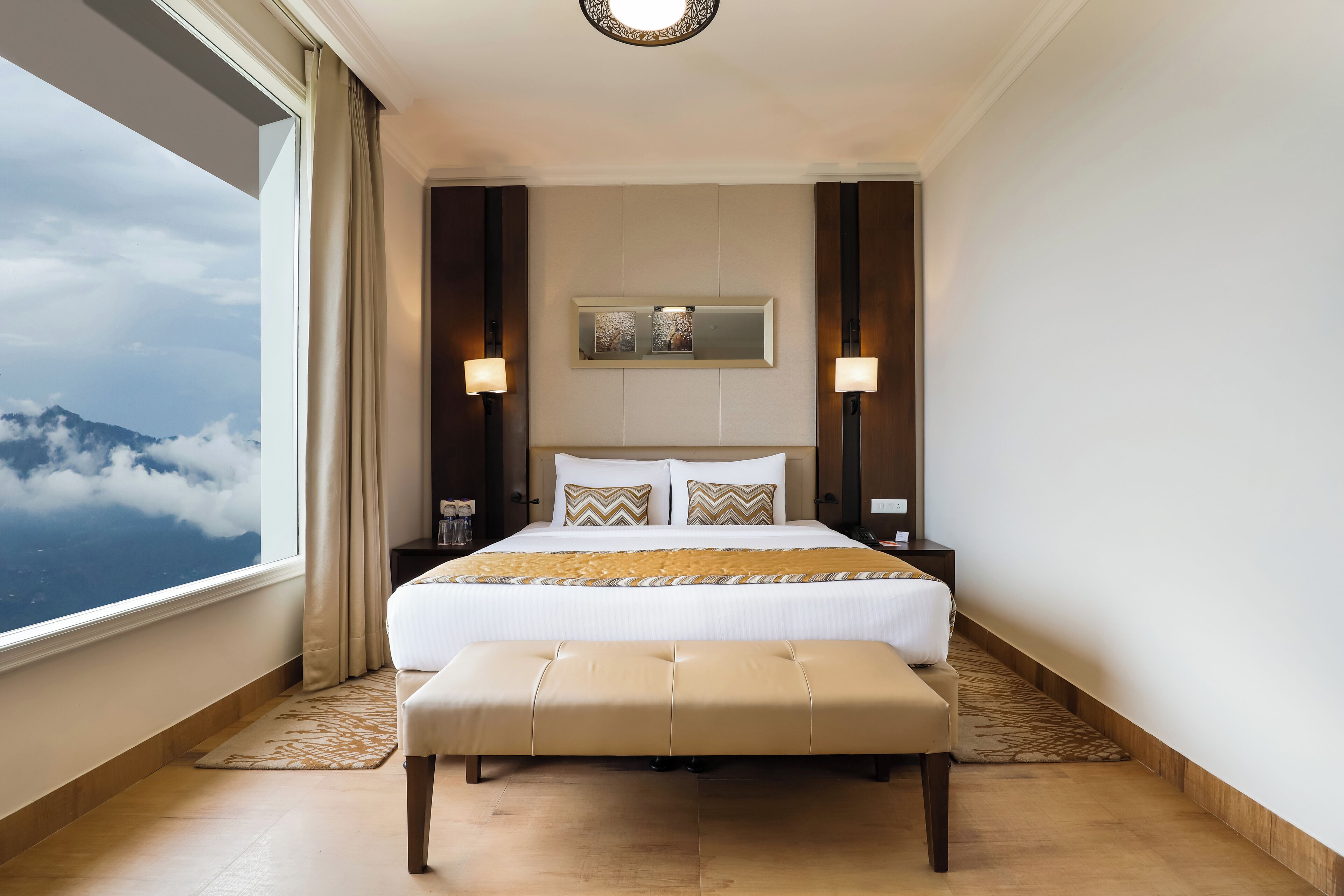 A hotel room with a large bed, a bench, a nightstand, and a view of clouds.