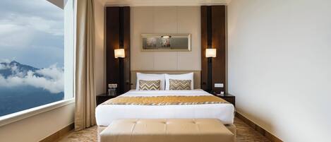 A hotel room with a large bed, a bench, a nightstand, and a view of clouds.