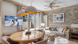Un salon moderne avec une table de salle à manger ronde en bois, des chaises rembourrées, un canapé, un pouf blanc et moelleux, un mur en pierre et un ventilateur de plafond.
