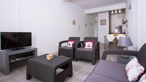 Een woonkamer met een flatscreen-televisie, twee rieten fauteuils met kussens, een bank, een salontafel en een keuken in de achtergrond.