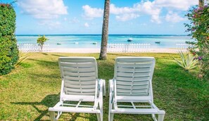 Two white lounge chairs facing a beach with clear blue water and a white fence in the background.