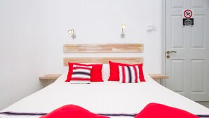Een net opgemaakt bed met rode en gestreepte kussens, een wit hoofdbord en een rookverbodsbord op de deur.