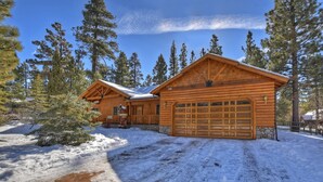 A log cabin with a large garage and a stone foundation, surrounded by snow and tall pine trees. - Stork and Bear Cabin by Destination Big Bear : Luxury Log Home in Eagle Mountain Estates! Pool Table! (Big Bear Lake)