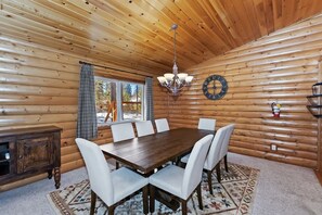 A wooden cabin interior with a dining table, white chairs, a chandelier, a clock, and a window with curtains.