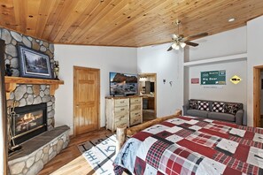 A bedroom with a stone fireplace, a bed with a red and white checkered bedspread, a television, and a ceiling fan.