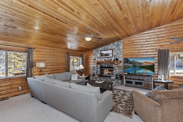 A log cabin interior with a stone fireplace, a large flat-screen TV, and a cozy seating area.