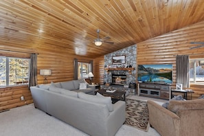 A log cabin interior with a stone fireplace, a large flat-screen TV, and a cozy seating area.