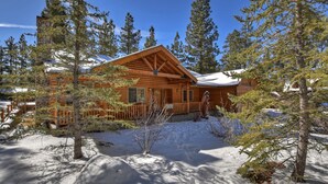 A log cabin with a stone chimney, surrounded by snow and tall pine trees. - Stork and Bear Cabin by Destination Big Bear : Luxury Log Home in Eagle Mountain Estates! Pool Table! (Big Bear Lake)