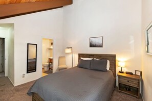 A bedroom with a large bed, a bedside table with a lamp, a framed picture on the wall, and a door leading to another room. - Snow Summit Chalet by Destination Big Bear : Next Door to Snow Summit for Easy Access to Skiing, Snowboarding and Tubing! (Big Bear Lake)