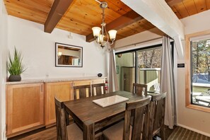 A dining area with wooden cabinets, a table, chairs, a chandelier, a potted plant, and a sliding glass door leading to an outdoor deck. - Snow Summit Chalet by Destination Big Bear : Next Door to Snow Summit for Easy Access to Skiing, Snowboarding and Tubing! (Big Bear Lake)