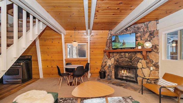 A cozy living room with a stone fireplace, a television mounted on the wall, a dining area with a table and chairs, and a staircase leading to another level. - Lower Moonridge! Close to Zoo, Bear Mountain Resort, Golf Course & Hiking/Biking Trails! Fenced Yard! (Big Bear Lake CA)
