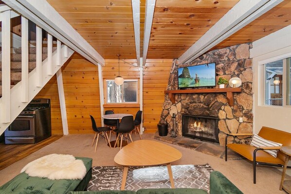 A cozy living room with a stone fireplace, wooden ceiling beams, a television, and a dining area.