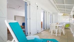 A sunlit room with a poolside chair, a dining table with chairs, and a door leading to another room.