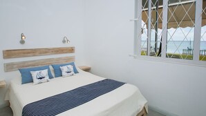 Un dormitorio con cama, dos mesitas de noche, ventana con vista al mar y cabecera de madera.