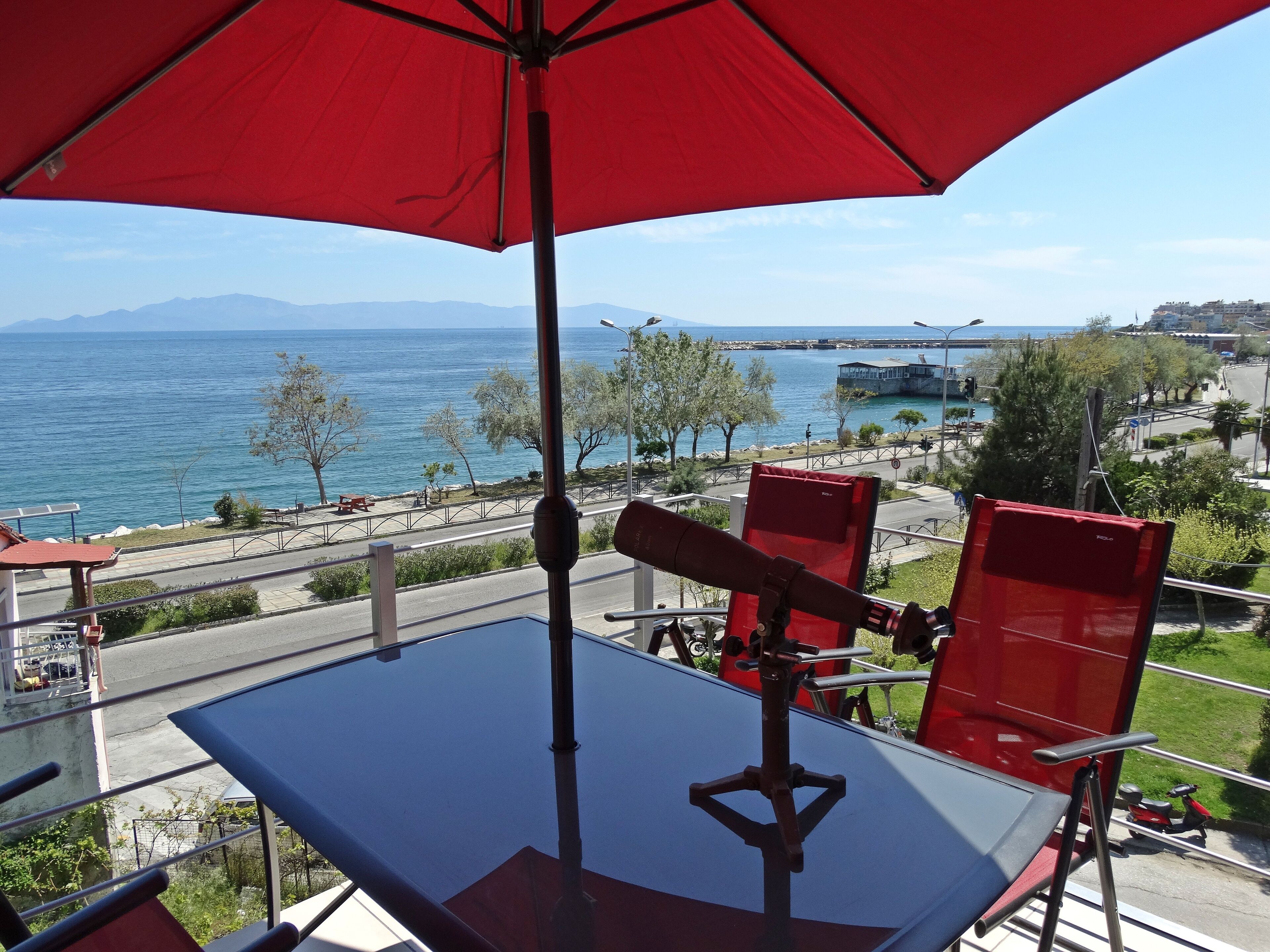 Varanda com um guarda-sol vermelho, uma mesa e cadeiras vermelhas, com vista para a costa, estrada e edifícios.