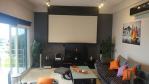 Ein modernes Wohnzimmer mit einem großen Flachbildfernseher, einer Couch mit gemusterten Kissen, einem Couchtisch und einer Schiebeglastür, die zu einem Außenbereich führt.