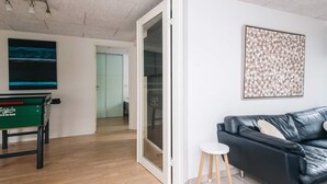 A modern living room with a black leather sofa, a small white stool, a green table with a logo, a glass door leading to another room, and a framed abstract art piece on the wall. - 5 Star Holiday Home in Henne (Henne)