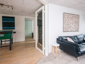 Ein modernes Wohnzimmer mit einer schwarzen Ledersofa, einem kleinen weißen Hocker, einem grünen Tisch mit einem Logo, einer Glastür zu einem weiteren Raum und einem gerahmten abstrakten Kunstwerk an der Wand.