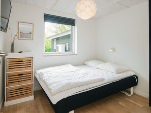 Une chambre à coucher comprenant un lit, une table de chevet, une commode avec des tiroirs, une fenêtre donnant sur un espace verdoyant et un luminaire suspendu.