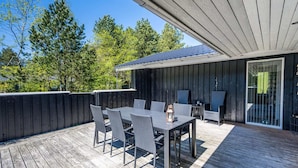 An outdoor deck with a dining table and chairs, a covered seating area, and a view of trees and a building in the background. - 4 Star Holiday Home in Norre Nebel (Norre Nebel)