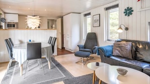 A modern living room with a sofa, a dining table with chairs, a kitchen area, and a large window. - 4 Star Holiday Home in Norre Nebel (Norre Nebel)