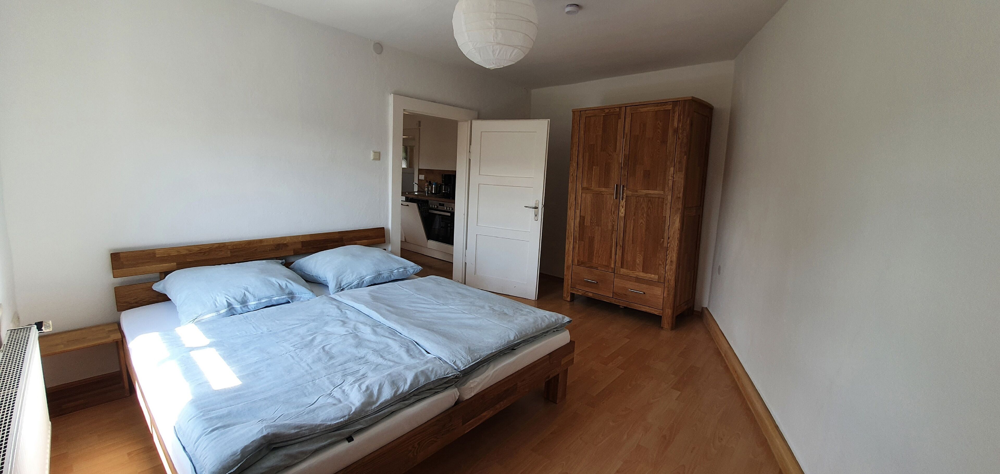 A bedroom with a wooden bed, a wardrobe, and a kitchen visible through an open door.