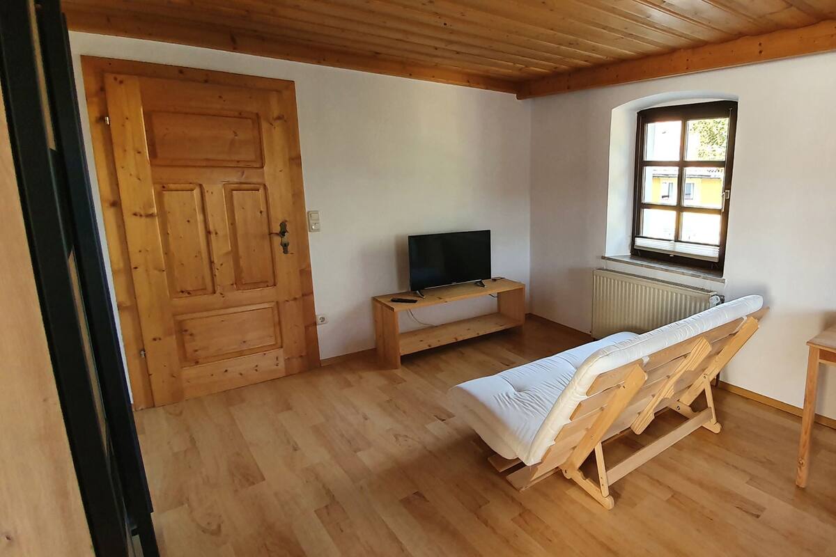 Ein Zimmer mit Holzboden, einem Bett mit weißer Matratze, einem Holztisch, einem Fernseher auf einem kleinen Tisch und einem Fenster mit Blick ins Freie.