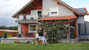 Una casa de dos pisos con un balcón, una antena parabólica y un rosal en el jardín delantero.