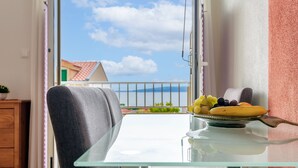 A balcony with a glass table and a chair, a plate of fruit, and a view of a building with a red roof.