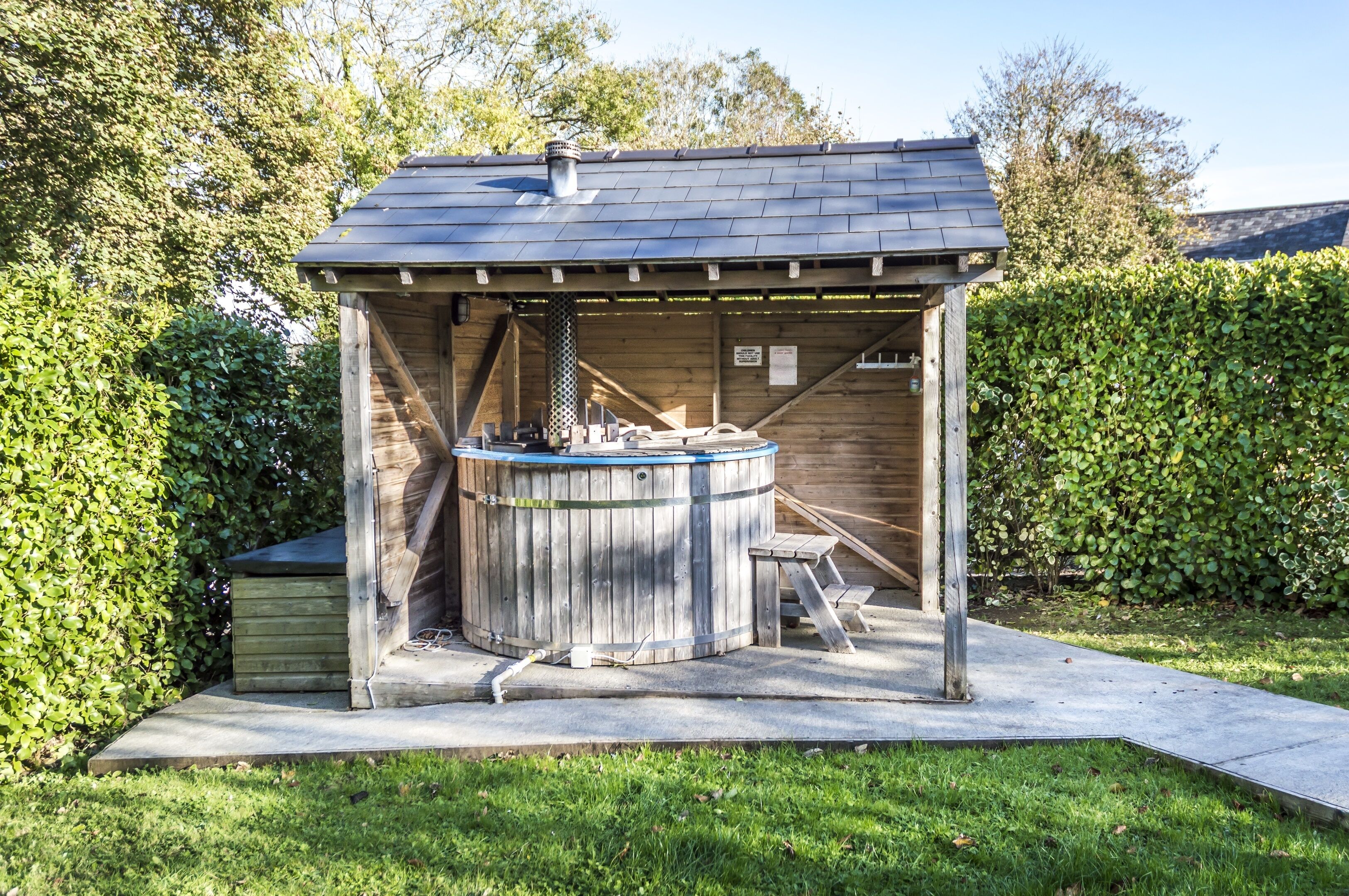A wooden outdoor hot tub structure with a metal roof, surrounded by a hedge and a grassy area.