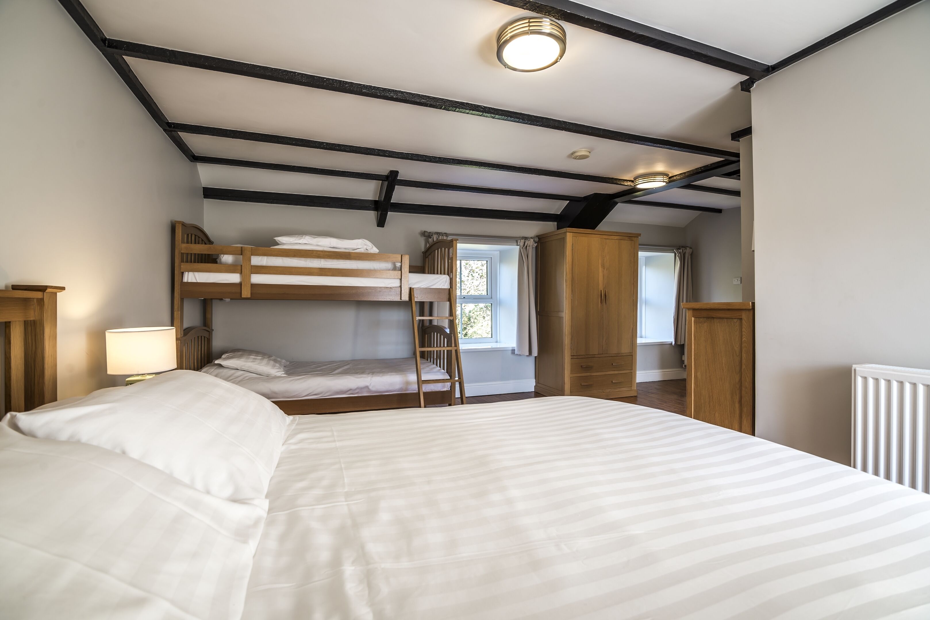 A bedroom with a large bed, a bunk bed, a wooden wardrobe, and a window with curtains.