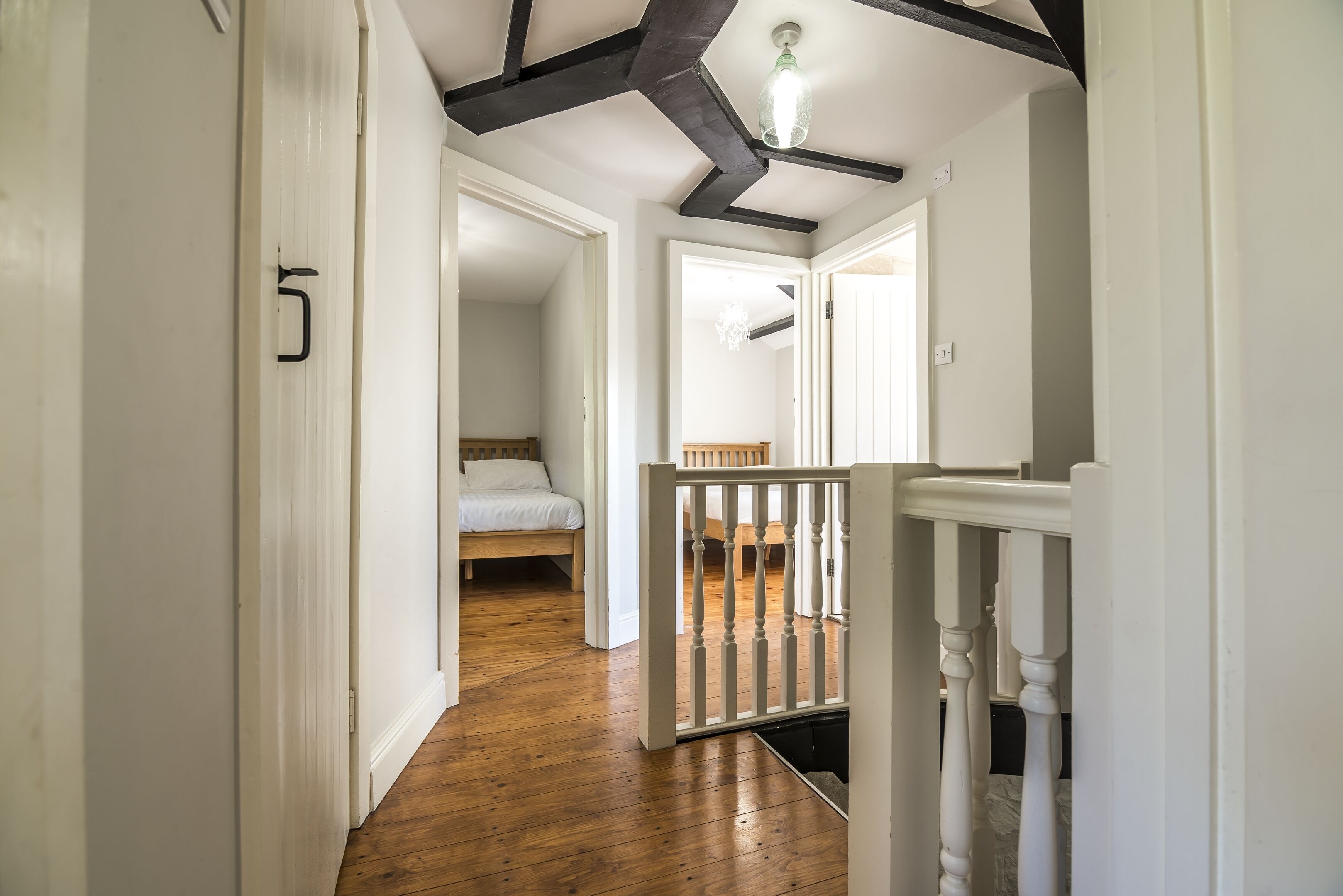 Un couloir avec un plancher en bois, des murs blancs et un plafond avec des poutres apparentes. On aperçoit un lit au fond.
