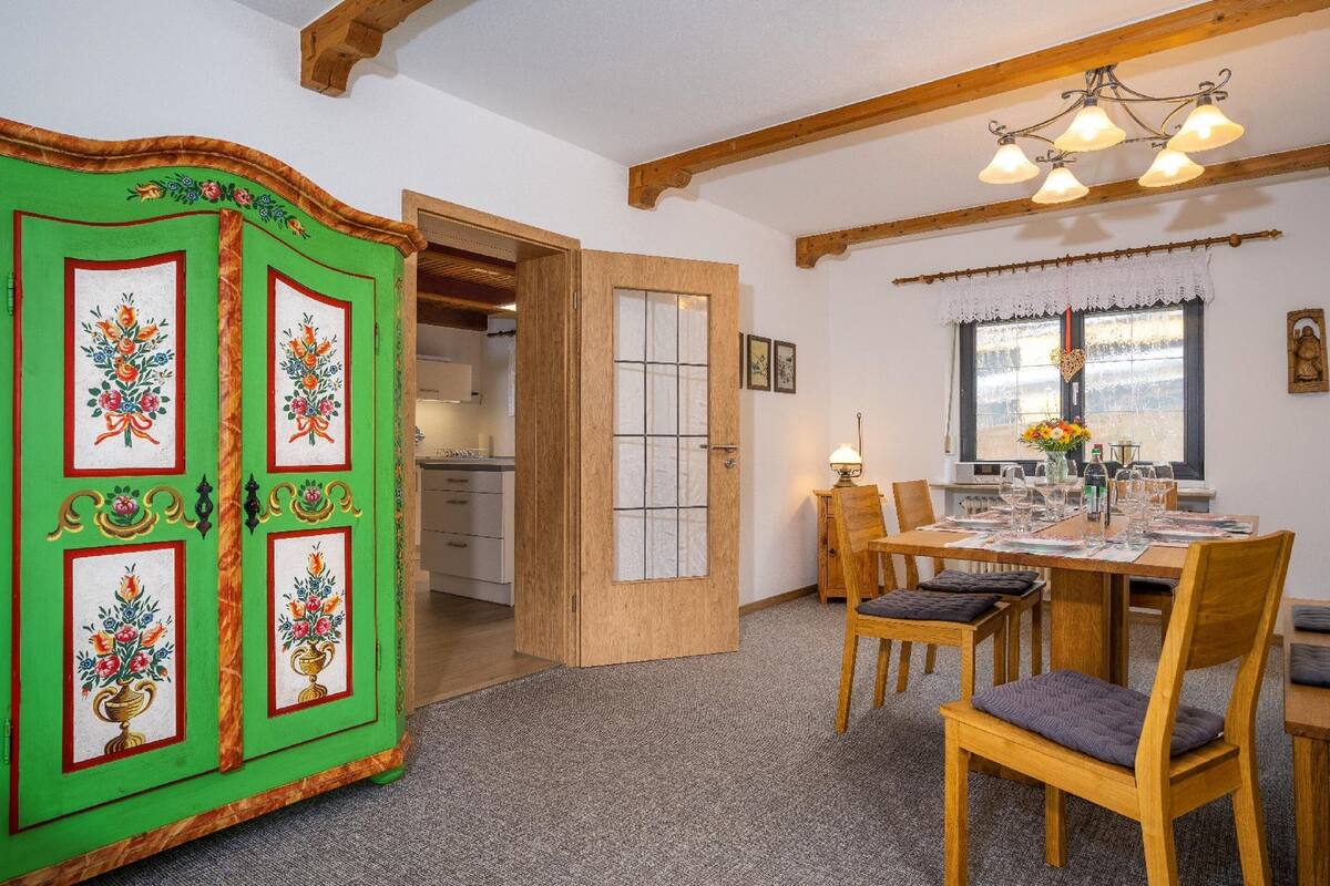 A dining area with a wooden table set for four, a green cabinet with floral designs, and a window with a lace curtain.