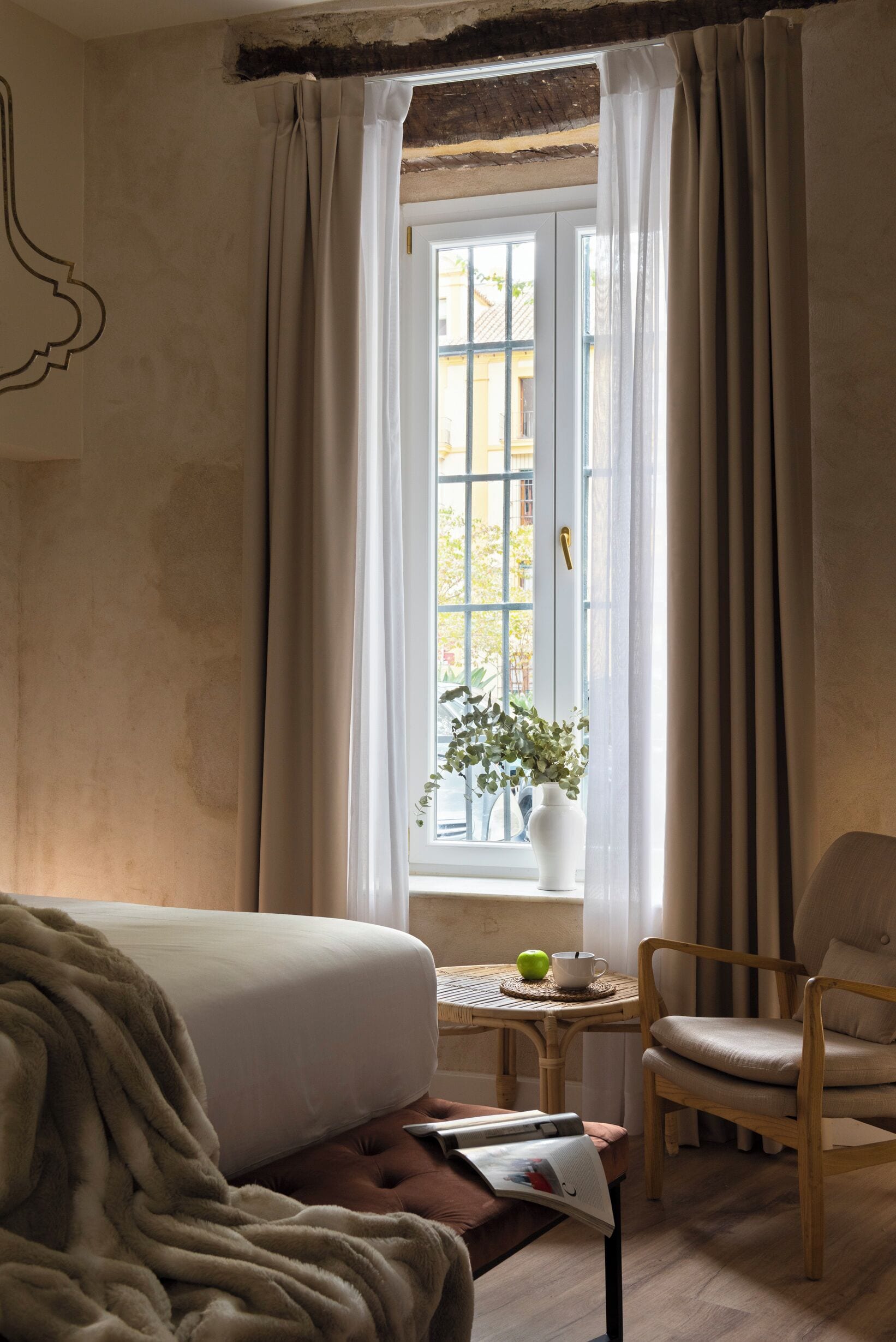 A hotel room with a bed, a chair, a small table with a plant, and a window with curtains.